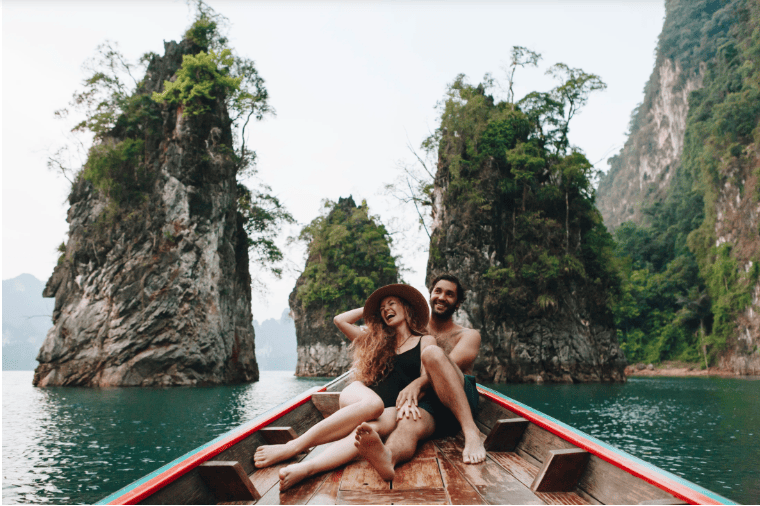 A couple relaxing on a wooden boat amidst lush limestone cliffs and clear blue water in Thailand, representing the country's focus on sustainable wellness and medical tourism