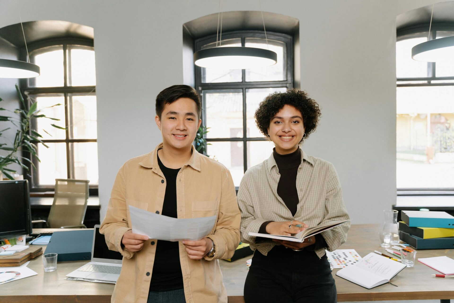 Two individuals in a professional office environment discussing documents. The background includes desks, office supplies, plants, and large windows, highlighting collaboration and productivity in a modern workspace.