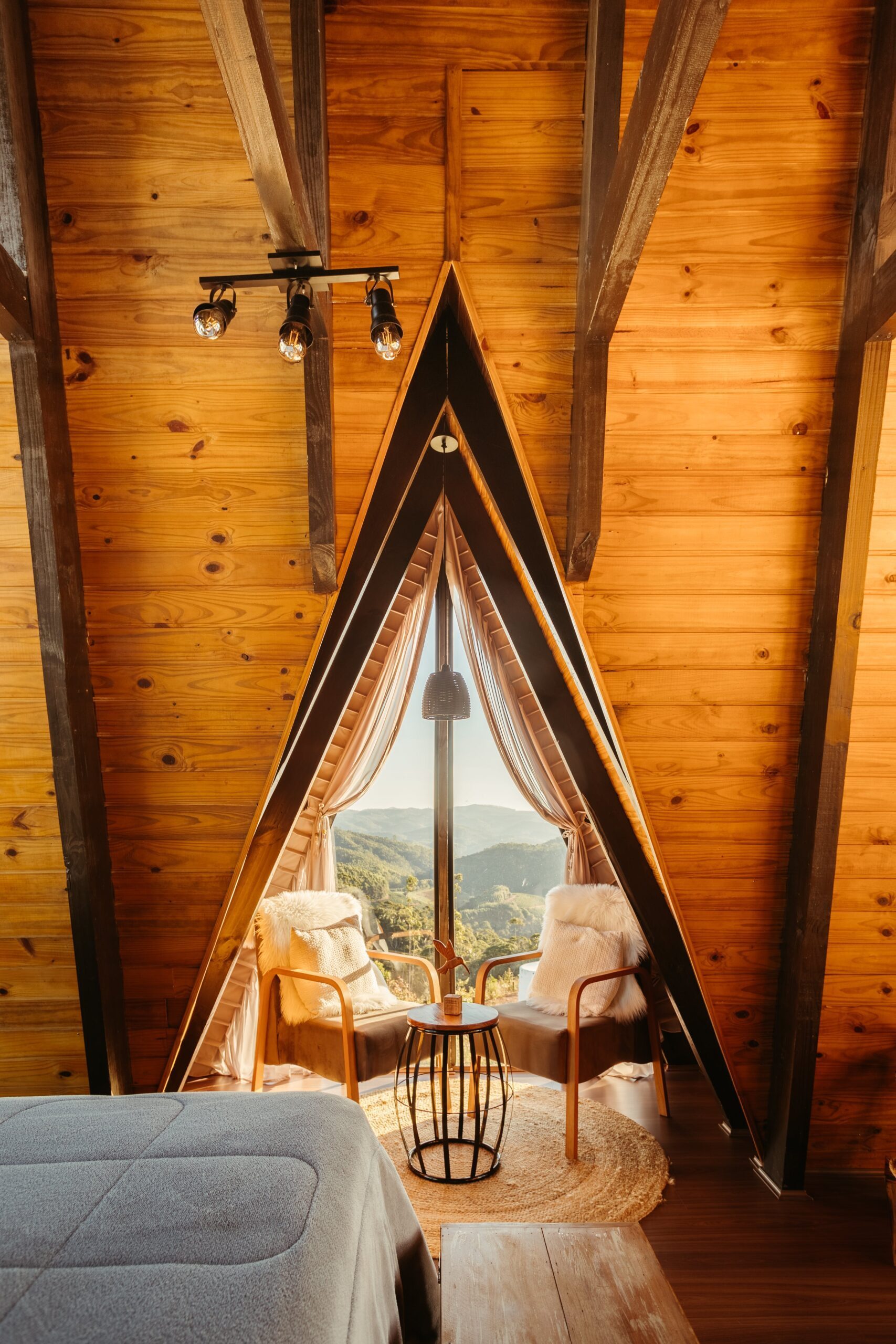A cozy, eco-friendly boutique hotel room with wooden interiors, featuring a triangular window with a scenic mountain view. The room includes two chairs with cushions, a small table, and a bed, emphasizing sustainable and inviting accommodations. This represents the REGENERA initiative to support boutique hotels with sustainable practices, eco-friendly operations, and enhanced guest experiences.