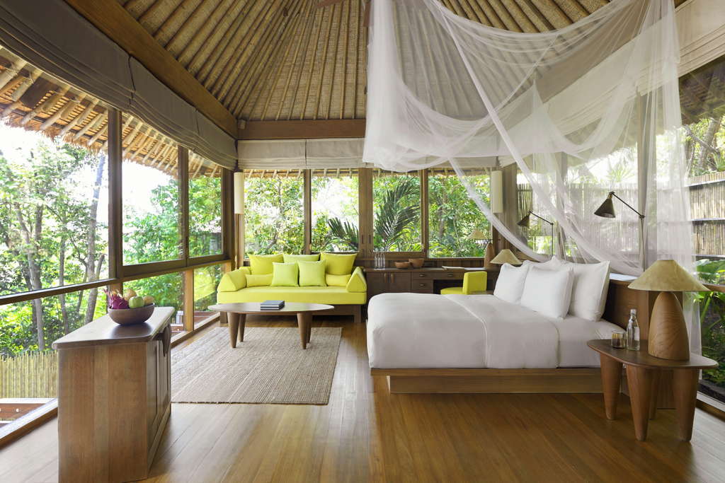 A boutique hotel room designed with eco-luxury principles, featuring a large bed with white linens and a mosquito net, tropical-themed furnishings, and large windows showcasing lush greenery. The serene setting reflects the REGENERA association's mission to promote regenerative sustainability, protect local communities, and support young talent in the boutique hotel industry.