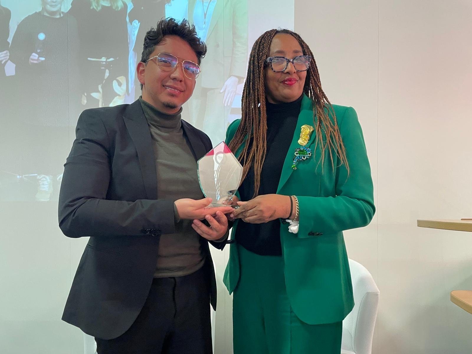 "Two representatives of Responsible Travel South America proudly holding a geometric-shaped award at a ceremony. They are standing in front of a large screen displaying their image, symbolizing recognition for their efforts in promoting gender equality across their business, fostering female leadership in tourism, and supporting women's roles in environmental initiatives.